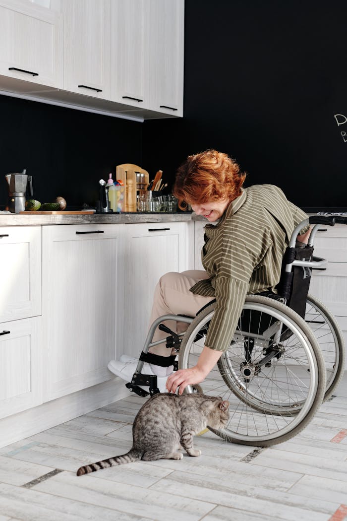 fitness-service-01 A woman in a wheelchair smiles while petting a tabby cat in a modern kitchen.